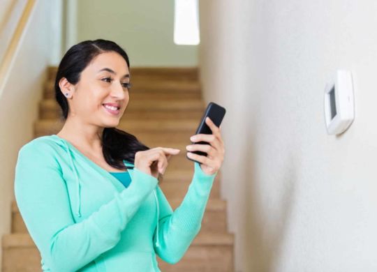 Pretty mid adult Hispanic woman uses smart phone to control temperature of home. She is standing in front of the thermostat on the wall in front of the stairs that lead to the second floor of her home. She smiles confidently as she uses the technology. She has brown hair pulled back into a pony tail and is wearing a light green hoodie.