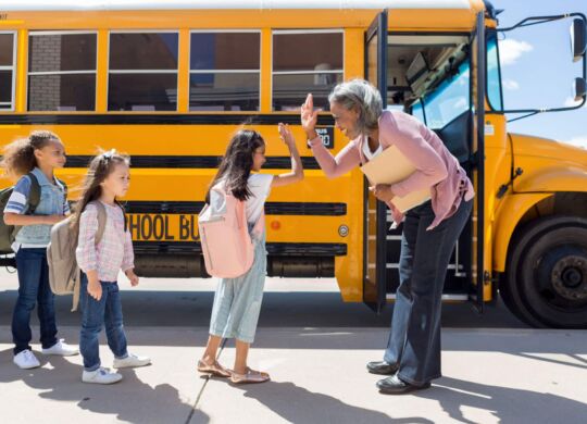 While waiting in line to board the school bus, a young female student gives the bus driver a high-five.