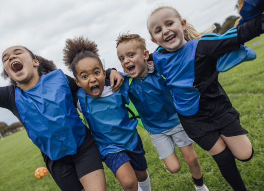 Front view of a small group of boys and girls wearing sports clothing, football boots and a sports bib on a football pitch in the North East of England. They are at football training where they are doing different football training drills. They are running and celebrating with their arms around each other while smiling and looking at the camera.

Videos are available for this scenario.